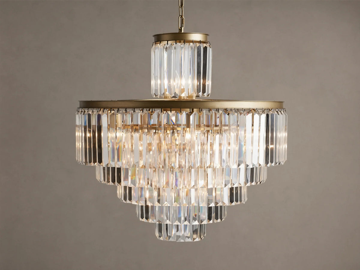 Leyland Chandelier, a tiered crystal fixture with clear rectangular prisms and an antique brass frame, glowing warmly against a plain wall.