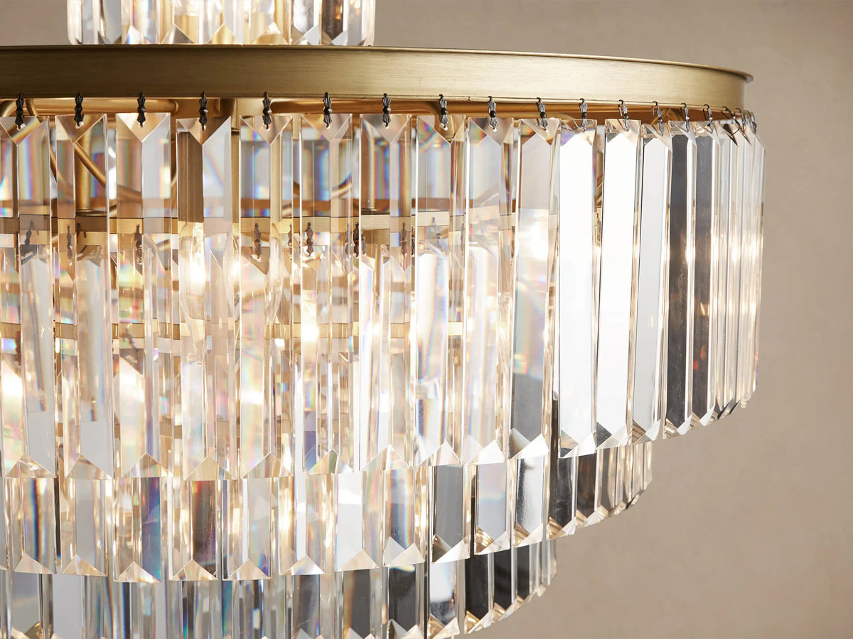 Leyland Chandelier, a tiered crystal fixture with clear rectangular prisms and an antique brass frame, glowing warmly against a plain wall.