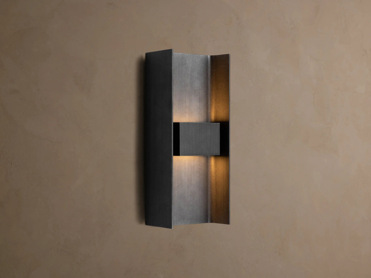 Sidney Outdoor Sconce, a graphite aluminum fixture casting warm LED light up and down against a taupe wall.