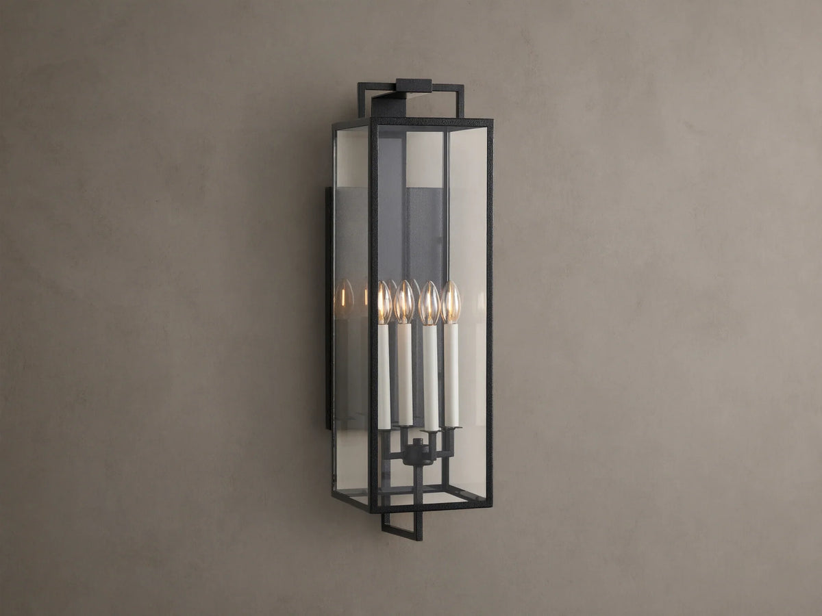 Hendricks Outdoor Sconce: black iron frame with clear glass panels, four lit candle-style bulbs, mounted vertically on a taupe wall.