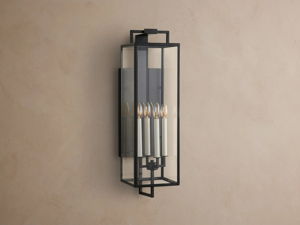Hendricks Outdoor Sconce: black iron frame with clear glass panels, four lit candle-style bulbs, mounted vertically on a taupe wall.