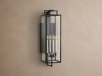 Hendricks Outdoor Sconce: black iron frame with clear glass panels, four lit candle-style bulbs, mounted vertically on a taupe wall.