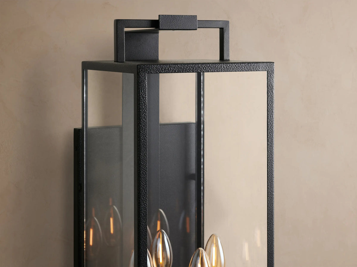 Hendricks Outdoor Sconce. Black iron rectangular lantern with clear glass panels, holding four white candle-style bulbs, mounted on a beige wall.