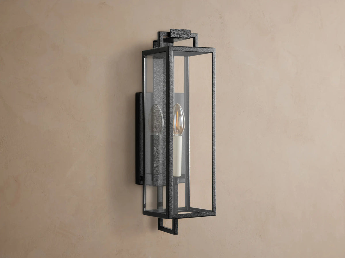 Hendricks Outdoor Sconce: dark iron rectangular lantern with clear glass panels, glowing candle-style bulb, mounted on a brown wall.