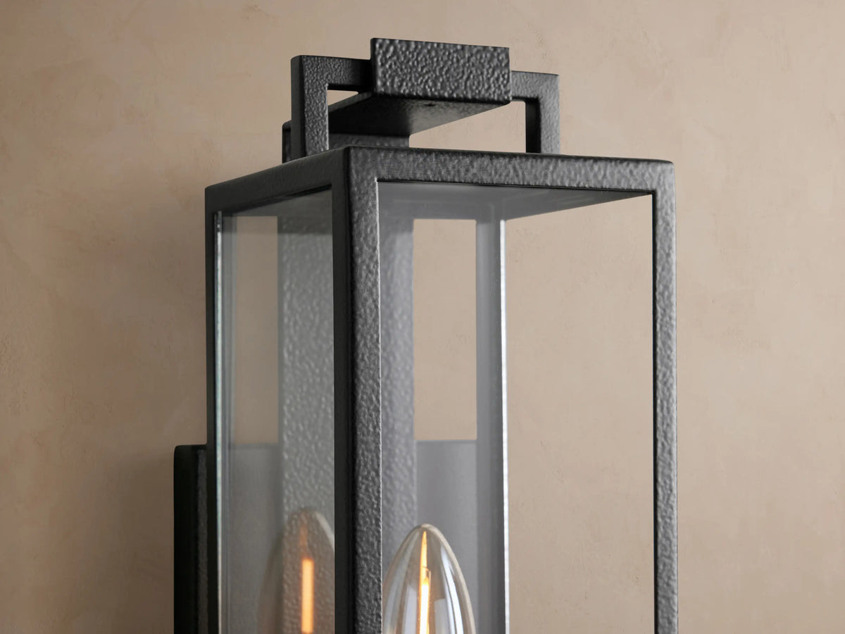 Hendricks Outdoor Sconce, black hand-worked iron and clear glass, rectangular, with one candelabra bulb, on a tan wall.