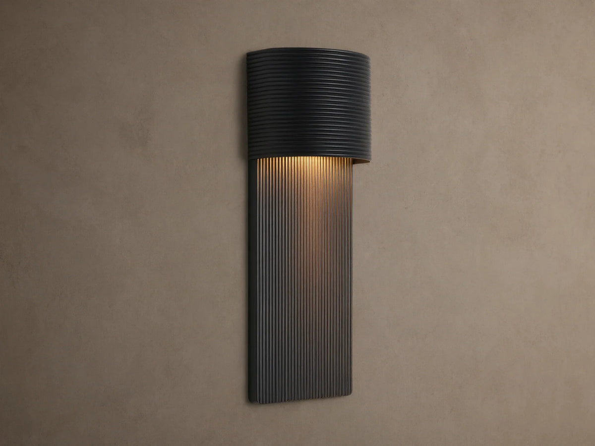 Fultz Outdoor Sconce, a soft black wall-mounted fixture with a horizontally ridged top and vertically ridged base, glowing warm light.