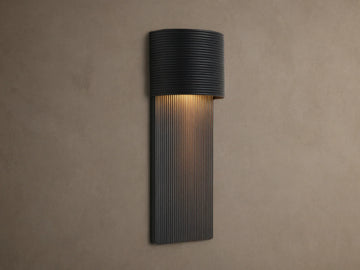 Fultz Outdoor Sconce, a soft black wall-mounted fixture with a horizontally ridged top and vertically ridged base, glowing warm light.