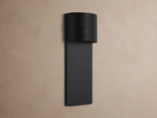 Fultz Outdoor Sconce, a soft black wall-mounted fixture with a horizontally ridged top and vertically ridged base, glowing warm light.
