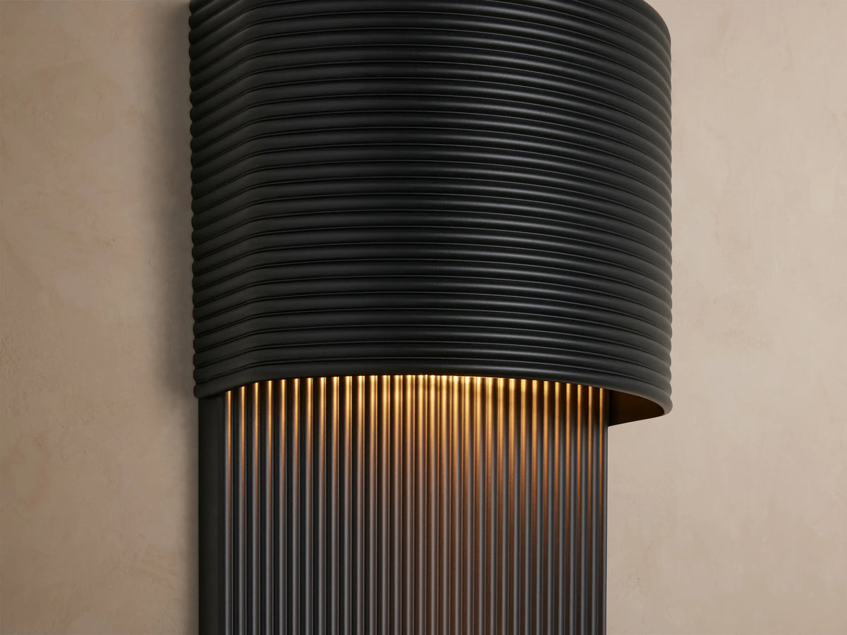 Fultz Outdoor Sconce, soft black, mounted vertically on a light beige wall, with a ribbed semi-circular top and ribbed rectangular bottom.
