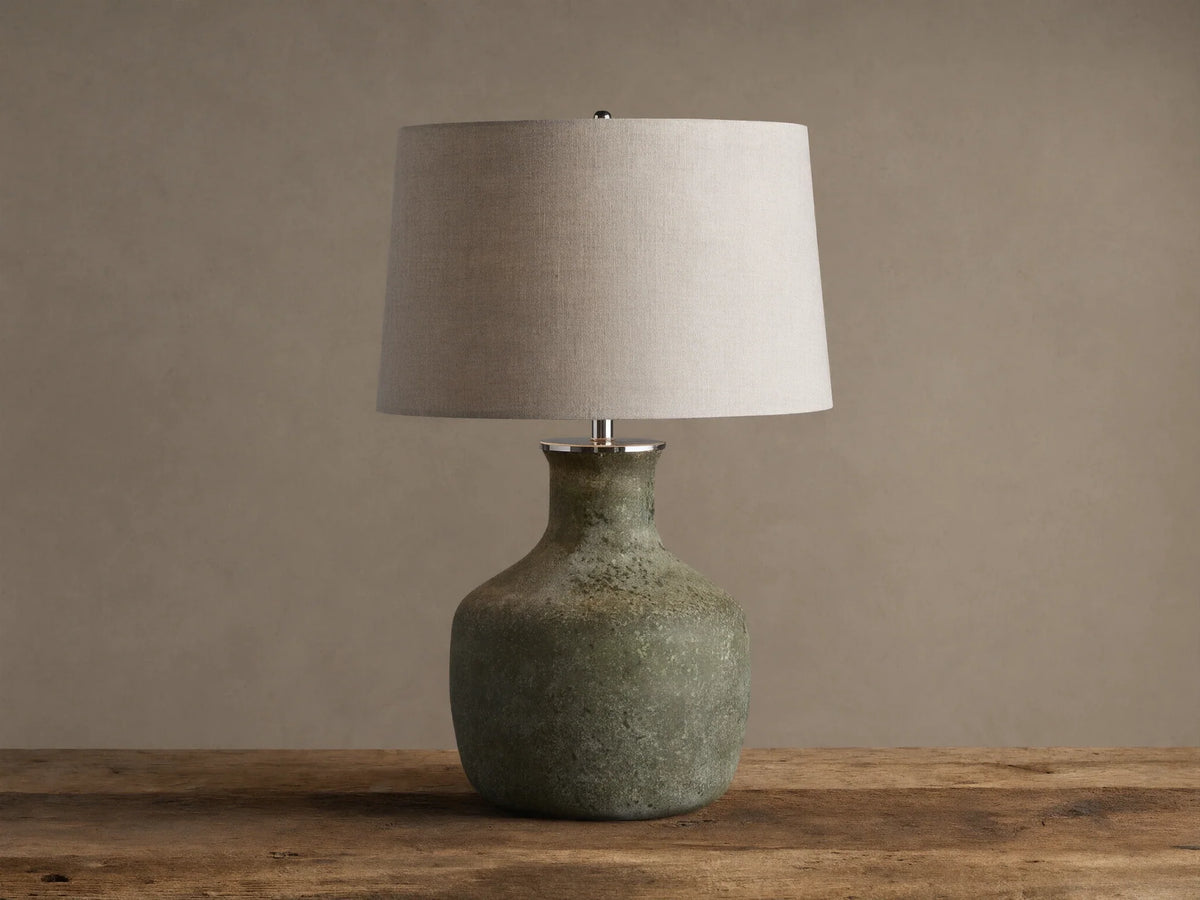 Galena Table Lamp: Green, sand-blasted glass bottle-shaped base with a light linen shade on a rustic wood surface.