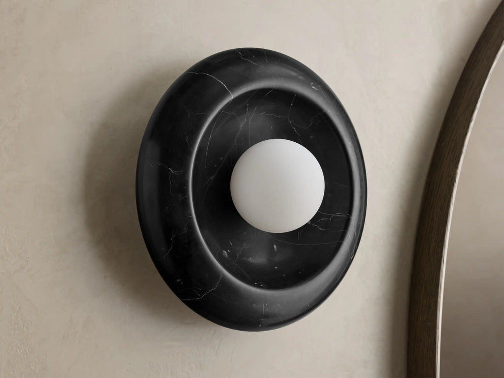 Front view of round black marble wall sconce with white spherical bulb, mounted on light wall next to wooden mirror frame.