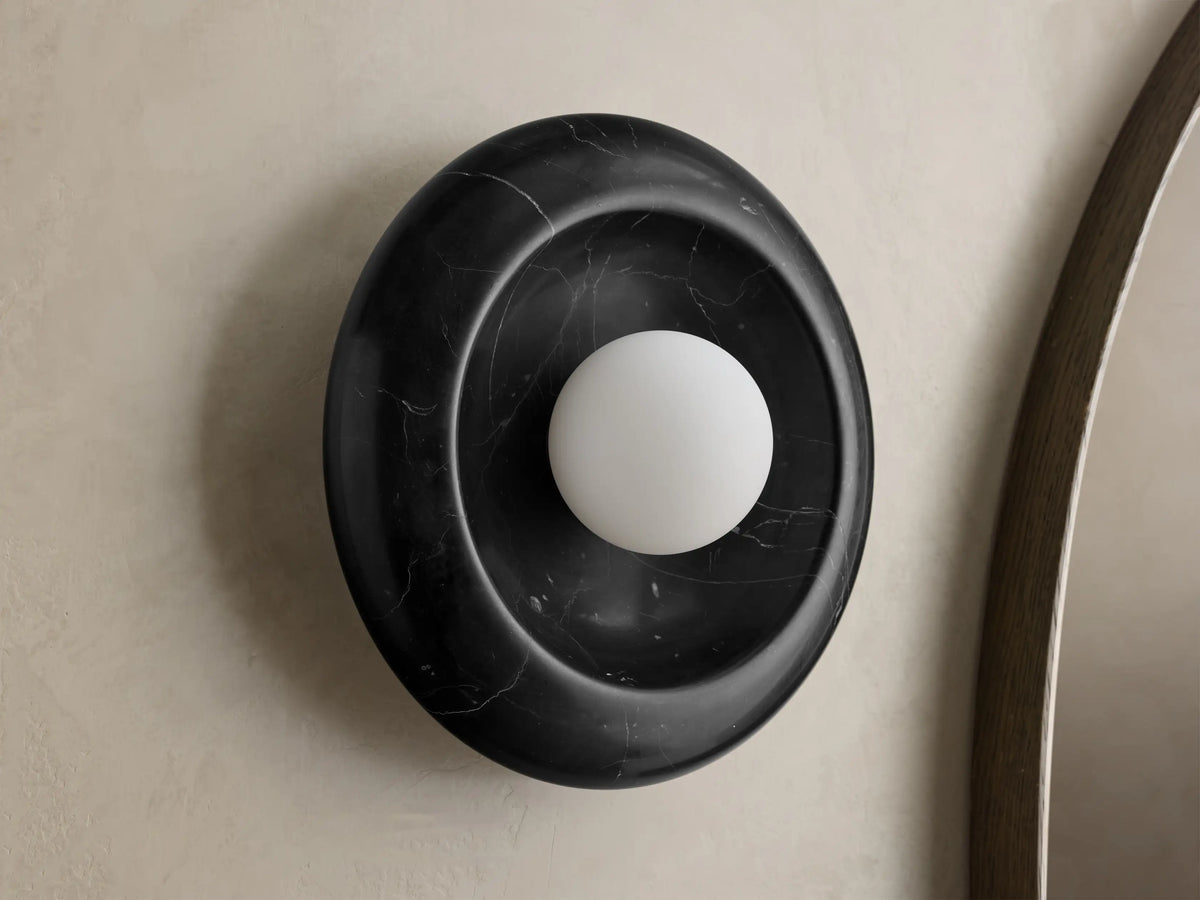 Front view of round black marble wall sconce with white spherical bulb, mounted on light wall next to wooden mirror frame.