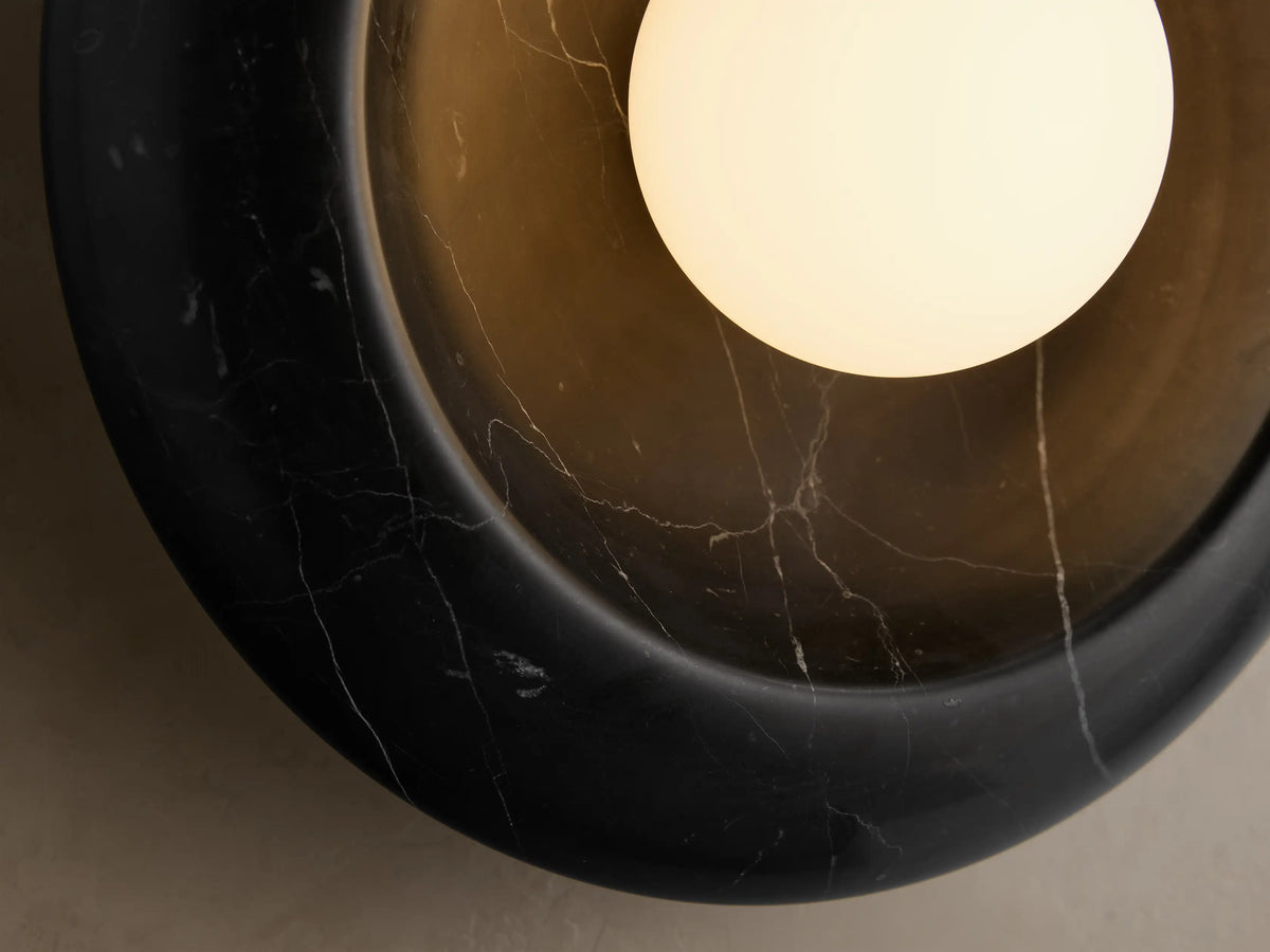 Close-up of a round black cracked marble wall sconce with a distressed gold interior and a glowing white spherical bulb.
