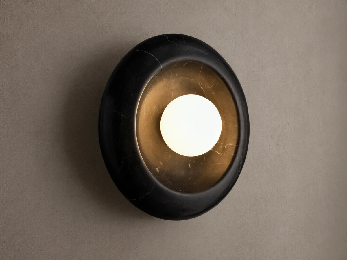 Close-up of a round black cracked marble wall sconce with a distressed gold interior and a glowing white spherical bulb.