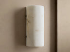 Wall-mounted cylindrical sconce made of alabaster, softly glowing with warm, inviting light against a beige textured wall.