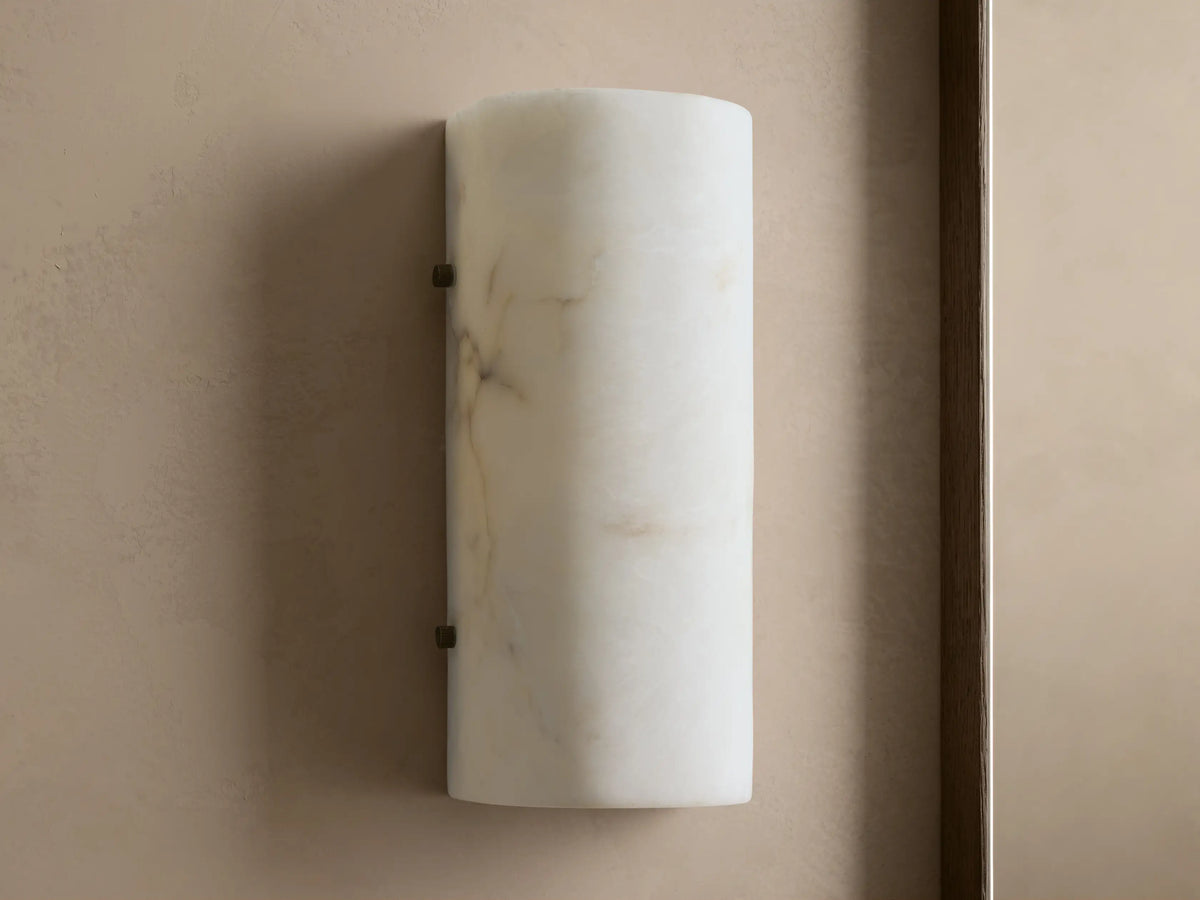 Wall-mounted cylindrical sconce made of alabaster, softly glowing with warm, inviting light against a beige textured wall.
