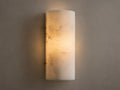Wall-mounted cylindrical sconce made of alabaster, softly glowing with warm, inviting light against a beige textured wall.