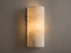 Wall-mounted cylindrical sconce made of alabaster, softly glowing with warm, inviting light against a beige textured wall.