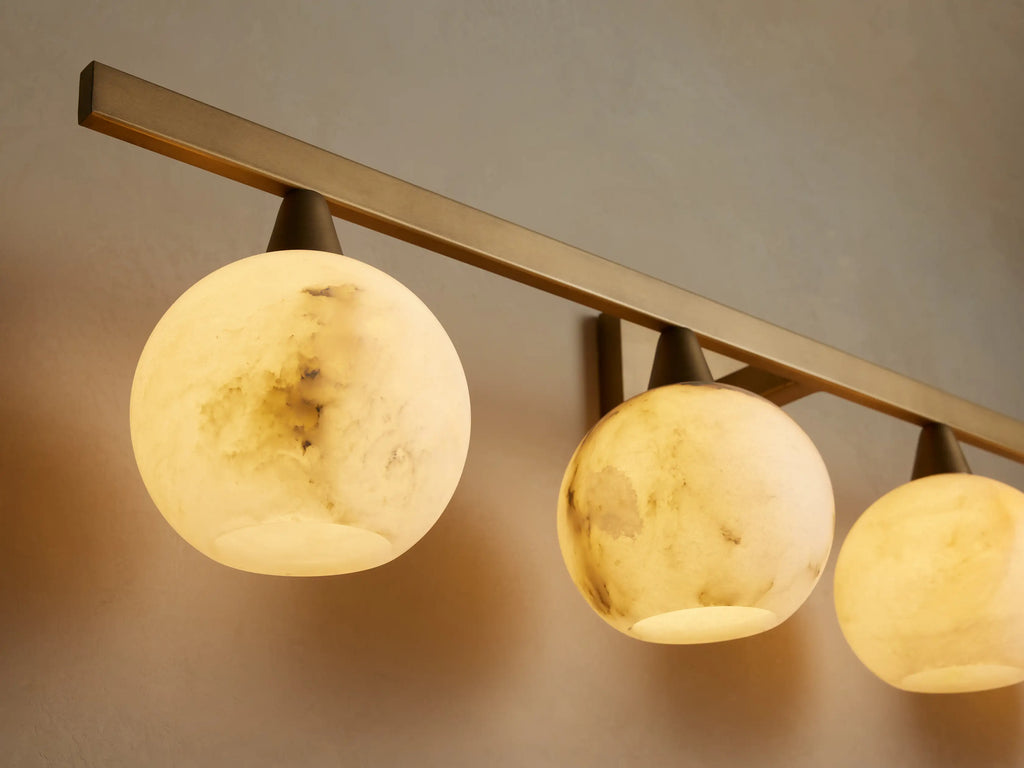 Arica Wall Sconce featuring a gold metal bar and four round white glass globes, mounted above a mirror on a beige wall.